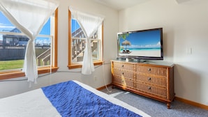 Una camera da letto con un letto, una televisione su una cassettiera in legno e due finestre con tende bianche.
