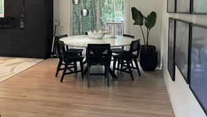 A modern kitchen with dark cabinetry, a round dining table with black chairs, and a large window with a view of greenery. - Minimalist sauna retreat  (Brighton)