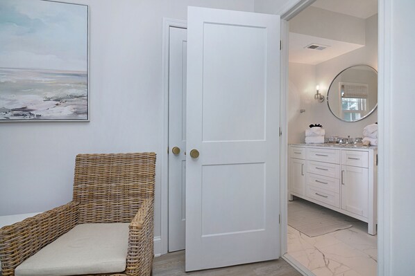 Una habitación con puerta blanca, una silla de mimbre y un baño con muebles blancos y un espejo.