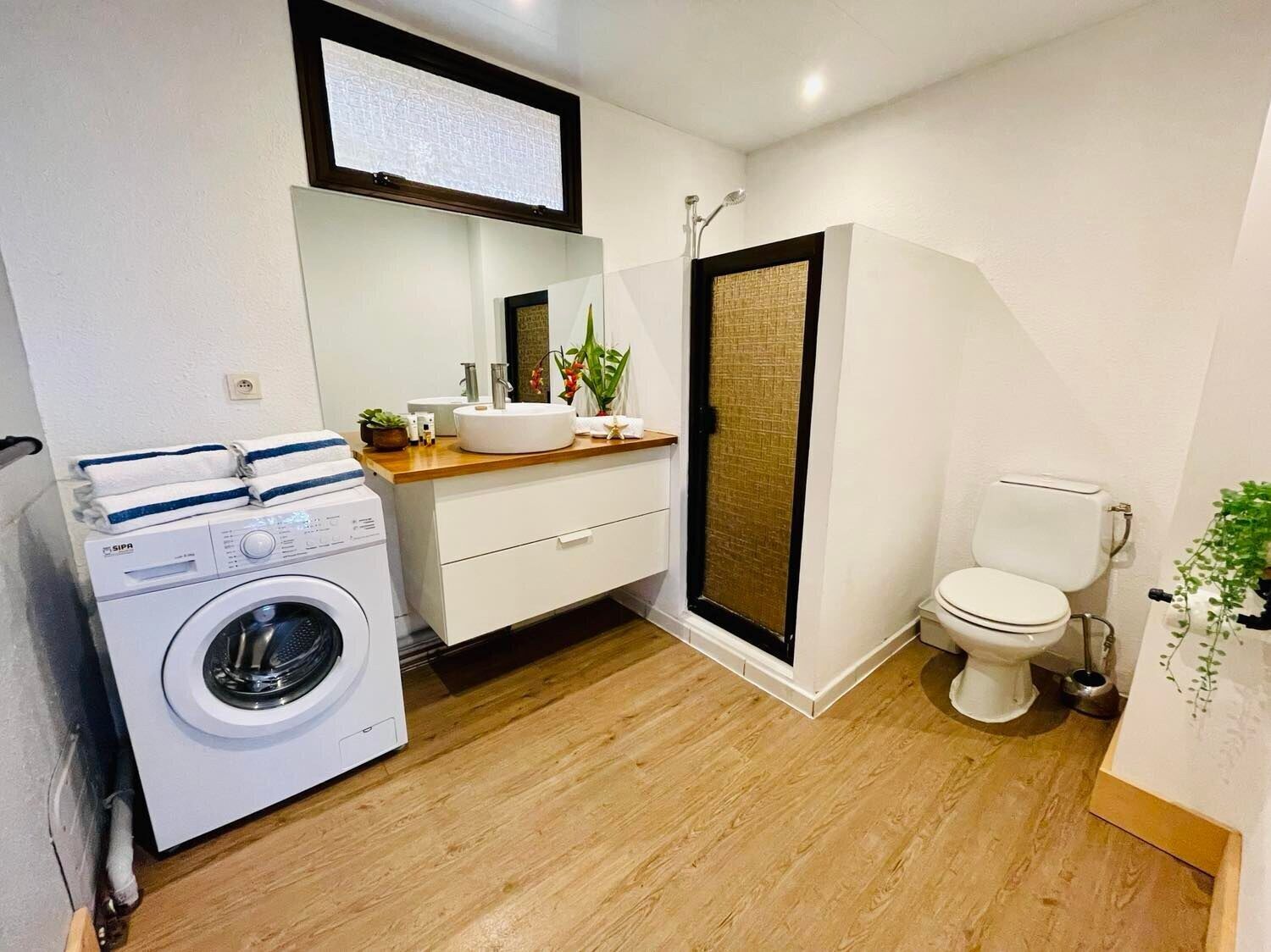 Et moderne badeværelse med vaskemaskine, vask med spejl, toilet og bruseområde.
