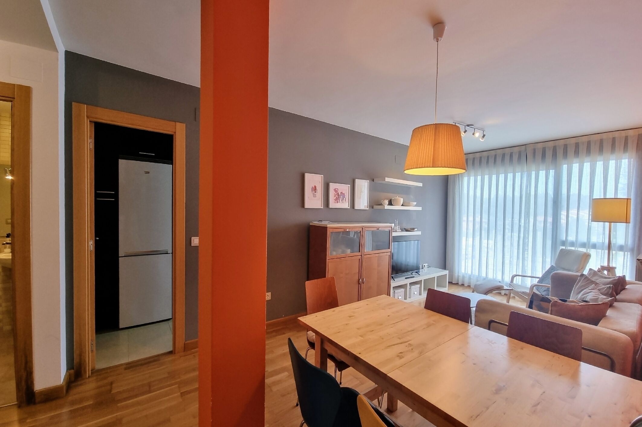 A modern living room with a wooden dining table, a sofa, and a TV. There is a kitchen area with a refrigerator and cabinets.