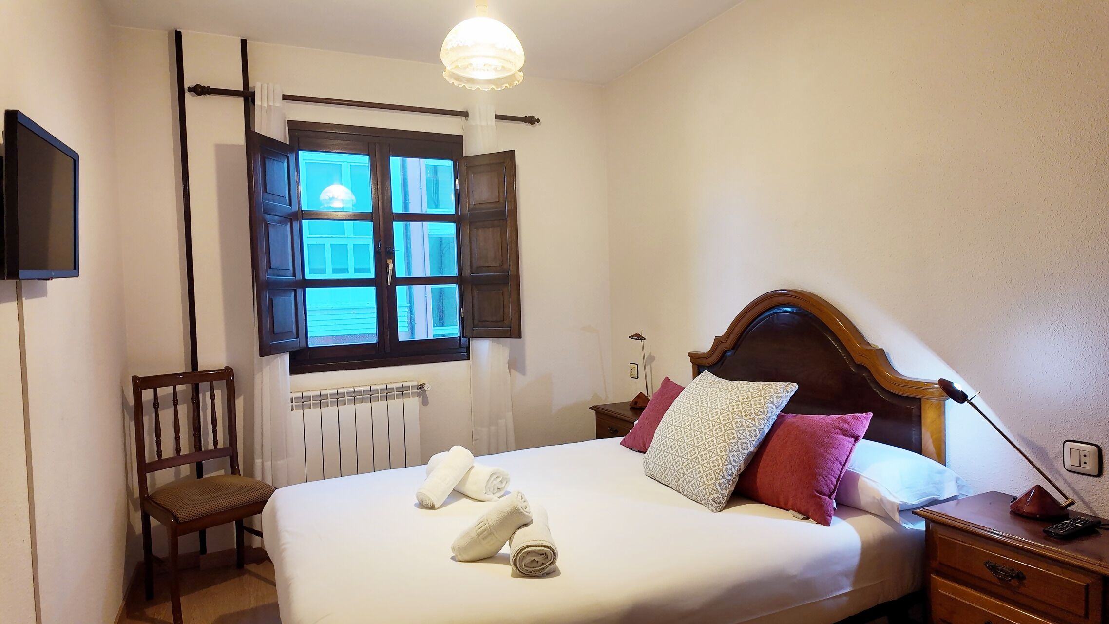 Ein ordentlich bezogenes Bett mit Kissen, ein Sessel, ein Nachttisch mit Lampe und ein Fenster mit Vorhängen.