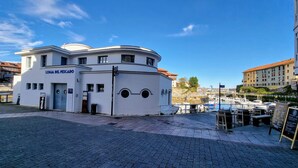 A white building with a dome roof labeled "LONJA DEL PESCADO" and a marina with boats in the background. - Apartamento Vacacional La Rula (Llanes)