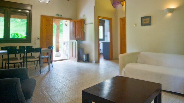 Amplia sala de estar con techo de madera, sofá blanco, mesa de comedor con sillas y área de cocina visible a través de un puerta abierta.