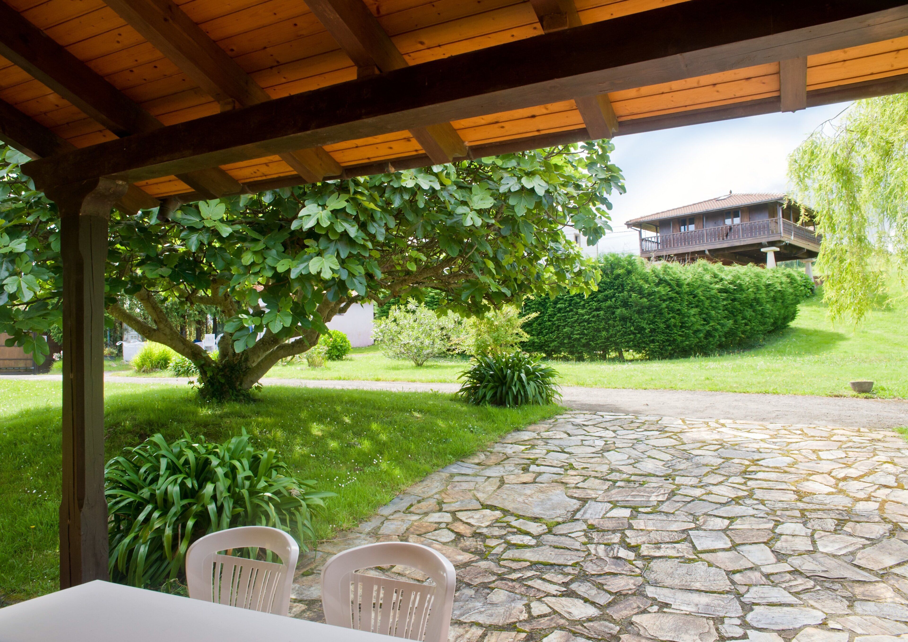 Un espace extérieur abrité, doté d’un sol en pierre, d’une table avec des chaises et d’un grand arbre qui offre de l’ombre.