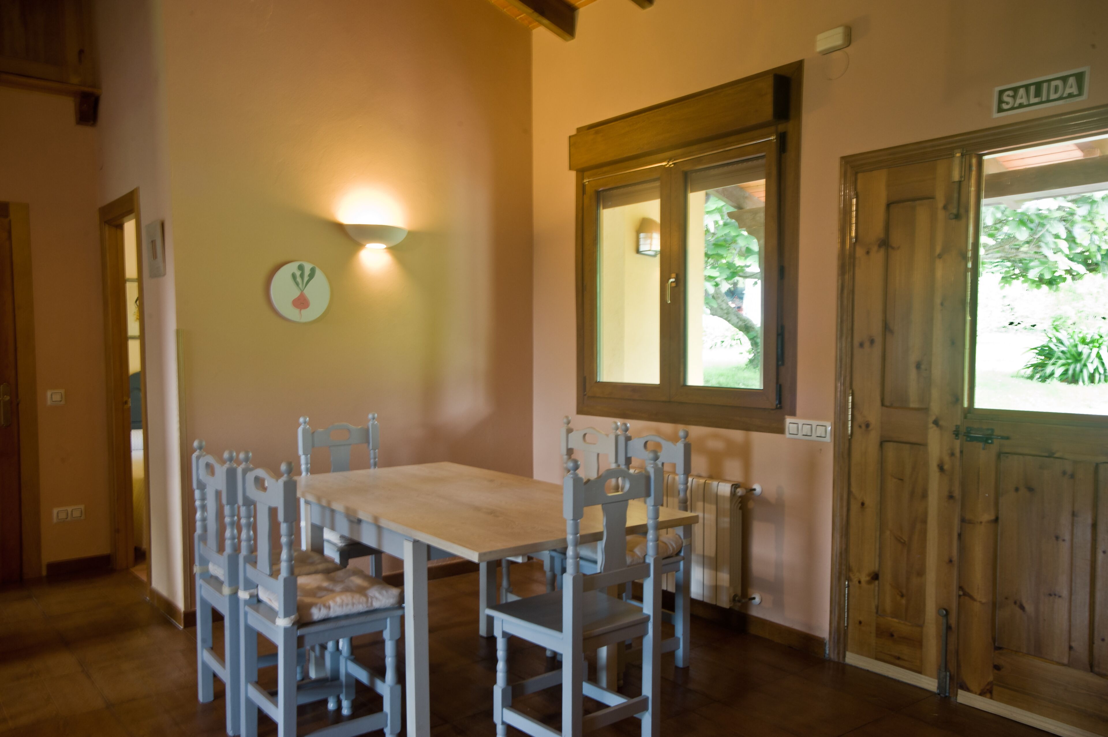 A dining area with a wooden table and chairs, a wall clock, a window with a view of greenery, and a door labeled 'SALIDA'.