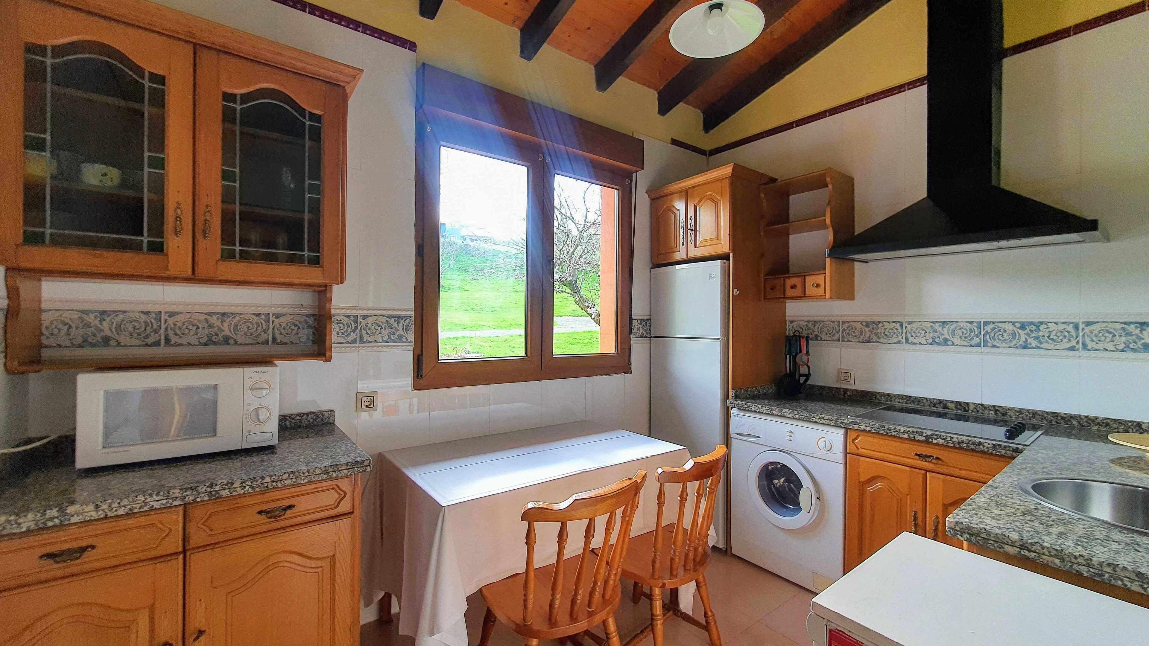 Een kleine keuken met houten kastjes, een magnetron, een wasmachine en een raam met uitzicht op groen.