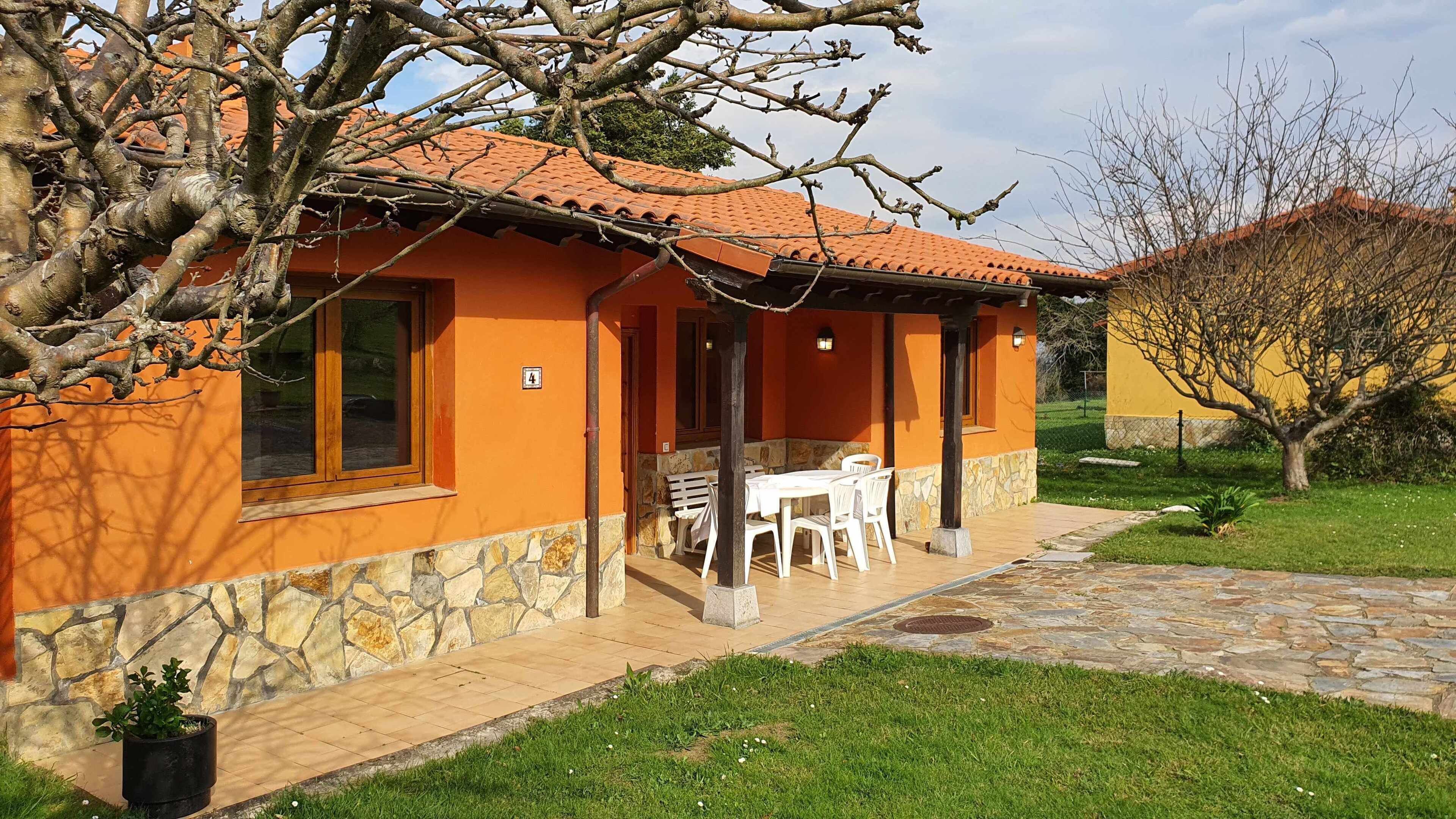 A house with a stone foundation, orange walls, and a tiled roof. There is a patio with a table and chairs, and a small garden area with a tree and grass.