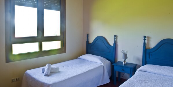 A room with two single beds, a nightstand with a lamp, and a window with blinds.