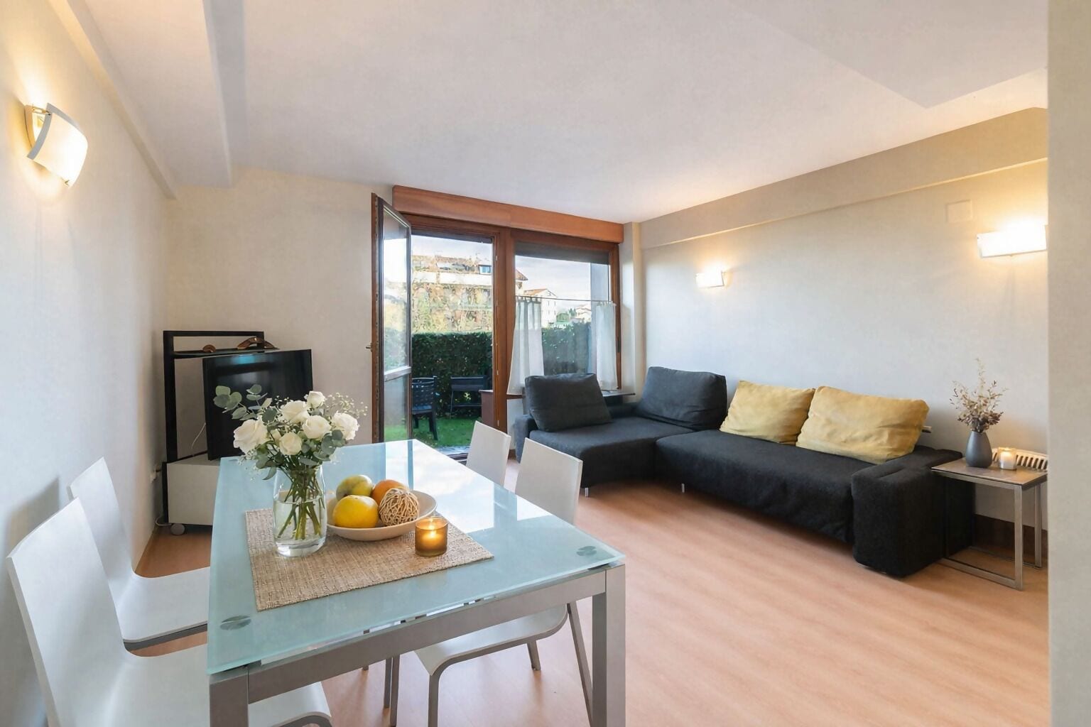 A modern living room with a glass dining table, a black sofa with yellow cushions, and a TV on a black stand.
