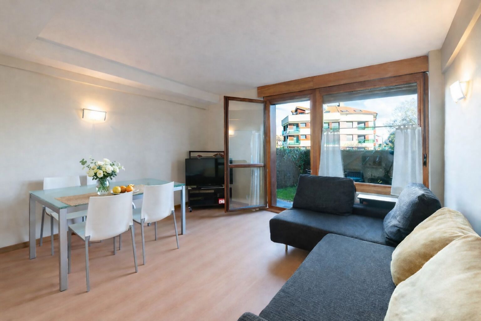 Ett modernt vardagsrum med en soffa, ett matbord med stolar, en tv och en skjutdörr i glas som leder ut till en balkong.