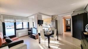 A modern living room with a sofa, a dining table with chairs, a kitchen area with a refrigerator, and a TV mounted on the wall. - La Terraza del Paraiso (Posada De Llanes)