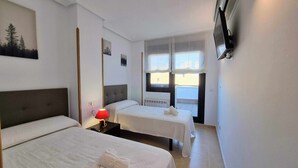 A hotel room with two beds, a red lamp, a TV, and a window with a view of buildings. - La Terraza del Paraiso (Posada De Llanes)