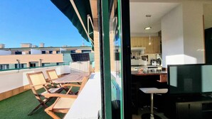 A balcony with wooden chairs and a table, a kitchen with wooden cabinets, and a view of neighboring buildings. - La Terraza del Paraiso (Posada De Llanes)