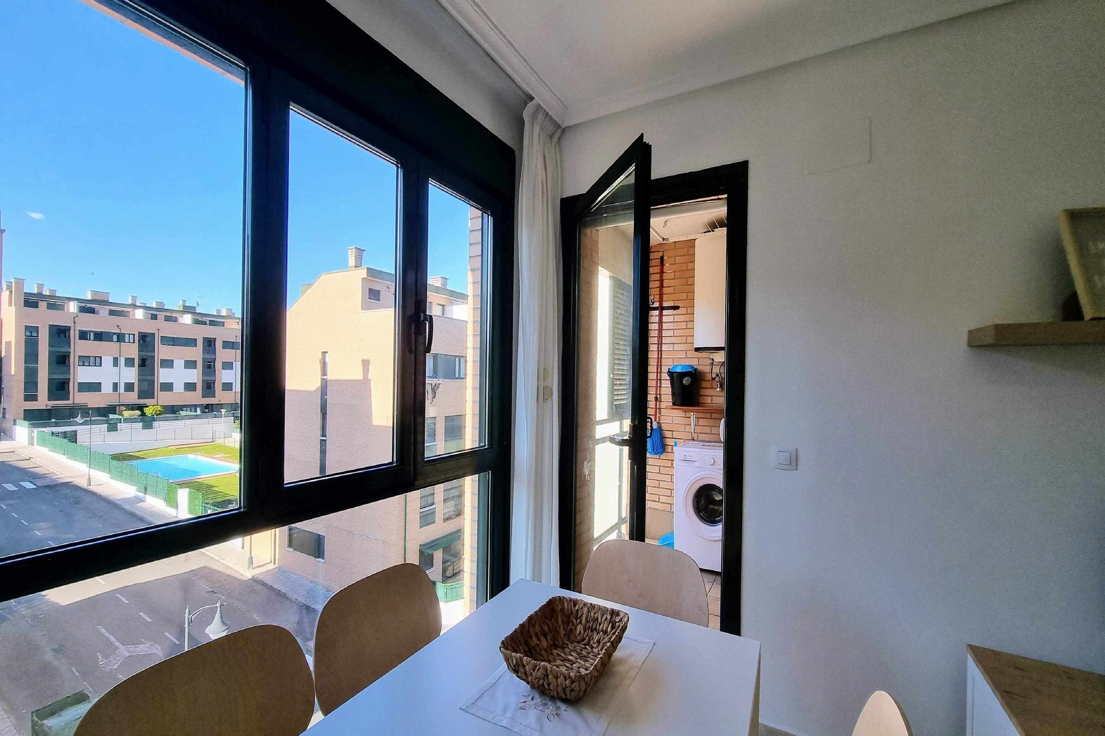 Une pièce dotée d’une grande fenêtre, d’une table sur laquelle se trouve un panier en osier, et qui offre une vue sur un bâtiment et une piscine situés à l’extérieur.