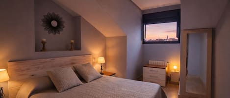 A modern bedroom with a large bed, bedside lamps, a nightstand, a dresser, a mirror, and a view of the city at sunset.