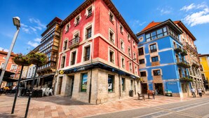 A red building with a blue and yellow facade, a street with outdoor seating, and a pedestrian walkway. - Apartamento Mercaderes (Llanes)