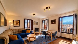 A living room with a sofa, a dining table, chairs, a TV, and large windows with a view. - Apartamento Mercaderes (Llanes)