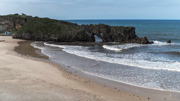 Een strand met een natuurlijke rotsformatie die over het water boogt, een zandstrand en een heldere lucht.