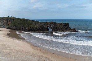 Une plage avec une formation rocheuse naturelle en forme d’arche surplombant l’eau, un rivage sablonneux et un ciel dégagé.