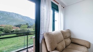 Een balkon met een beige bank, uitzicht op een groen landschap en bergen, en een wit gordijn.