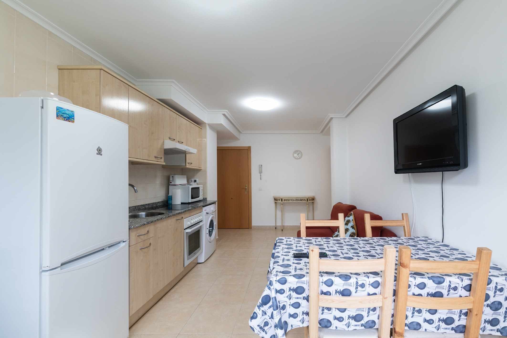 A compact kitchen with a refrigerator, microwave, oven, and washing machine. There is a dining area with a table and chairs, and a television mounted on the wall.