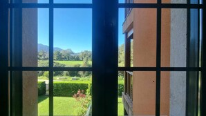 Une vue à travers une fenêtre à carreaux, dévoilant un paysage verdoyant avec une haie, un ciel dégagé et des montagnes lointaines.