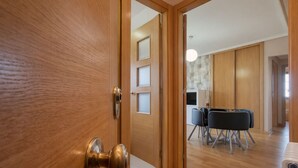 Une porte en bois dotée d’une poignée en laiton donne sur une pièce où se trouvent une table à manger et des chaises, un radiateur et un luminaire au plafond.