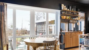 Un espace repas avec une table ronde et des chaises, une cuisine dotée de meubles et d’étagères en bois, et de grandes fenêtres offrant une vue sur l’extérieur.