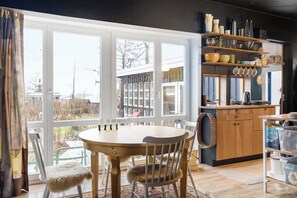 Un comedor con una mesa redonda y sillas, una cocina con armarios y estantes de madera, y ventanas amplias con vistas al exterior.