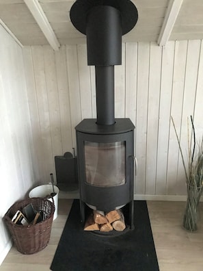Un poêle à bois doté d’une porte en verre, un panier de bois de chauffage et un seau blanc avec une louche.