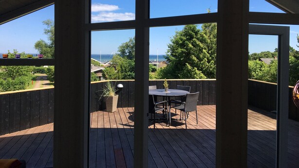 A covered outdoor deck with a table and chairs, a potted plant, and a view of the ocean and greenery. - Cottage in Grønninghoved with view (Sjølund)
