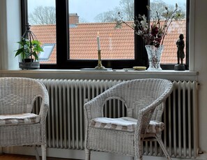 A room with two wicker chairs, a radiator, a window with a view of a red-tiled roof, a potted plant, a candle, and a vase with flowers. - Charming holiday apartment in central Stege (Stege)