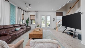 A living room with a brown leather sofa, a white armchair, a woven ottoman, and a flat-screen TV on a stand.