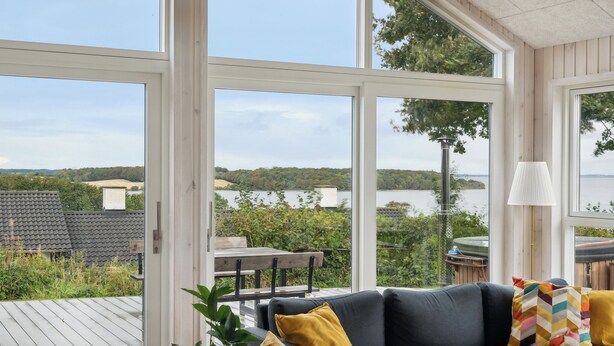 A modern interior with large windows, a grey sofa, and a view of a lake and greenery. - Lovely, new-built cottage near sea (Aabenraa)