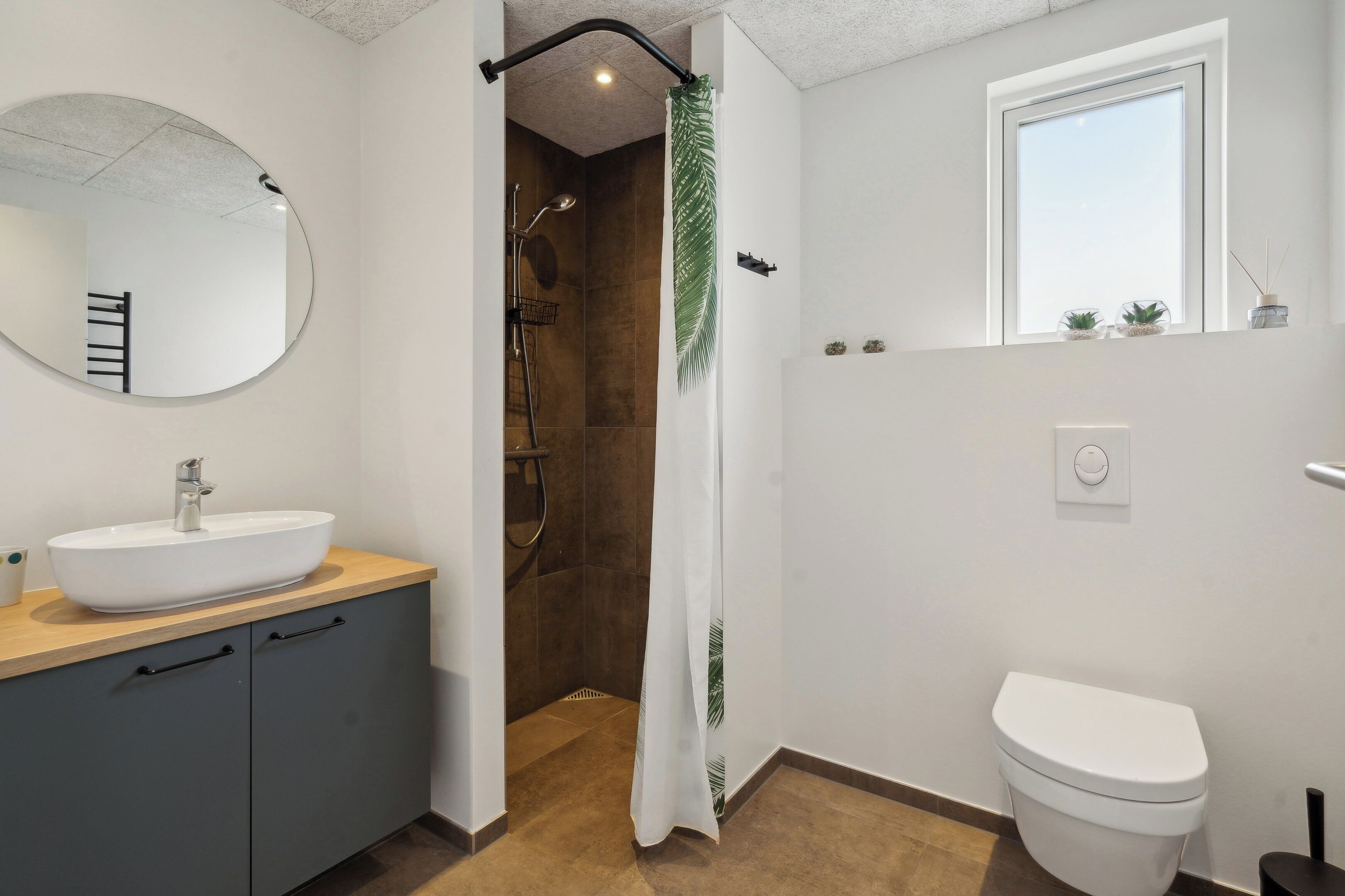 Un bagno con un lavandino bianco, mobili scuri, una doccia con tenda a foglia di palma, un WC e una finestra.