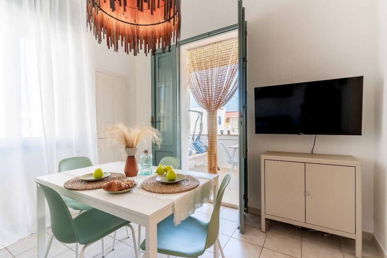 Un comedor con una mesa blanca, sillas verdes, un televisor y un mueble color beige.