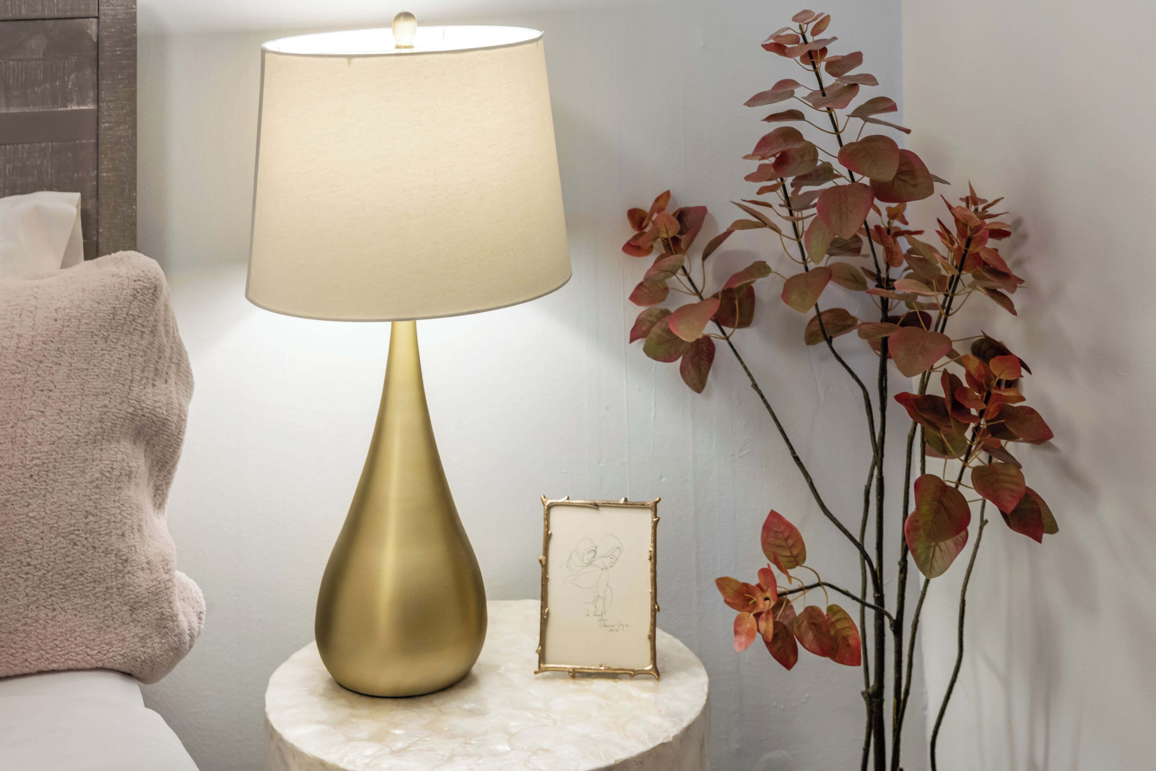 Ein Nachttisch mit einer goldenen Lampe, einem gerahmten Bild und einer Pflanze mit roten Blättern.