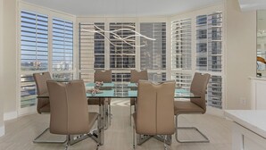 Un área de comedor con una mesa de vidrio y sillas marrones, ventanas amplias con persianas y vistas a edificios.