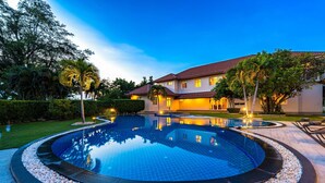 A large, circular swimming pool with a pebble border, surrounded by a well-manicured lawn and palm trees. - Luxury 6 Bed Mansion - CAS (Cha-am)