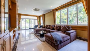 A spacious living room with a large sectional sofa, a flat-screen TV, and a large window with curtains. - Luxury 6 Bed Mansion - CAS (Cha-am)