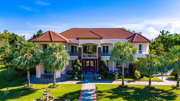 A large, two-story house with a red-tiled roof, a central entrance with a staircase, and a well-maintained lawn with palm trees. - Luxury 6 Bed Mansion - CAS (Cha-am)