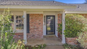 A two-story brick house with a front porch, a black door, and a small garden area.