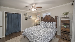A bedroom with a bed, a ceiling fan, a nightstand, a lamp, a wall clock, and a window with curtains.