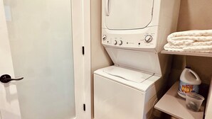 Une buanderie équipée d’un lave-linge et d’un sèche-linge à chargement frontal, d’une petite étagère avec une bouteille de lessive et un gobelet en plastique, et d’une porte dotée d’une poignée.