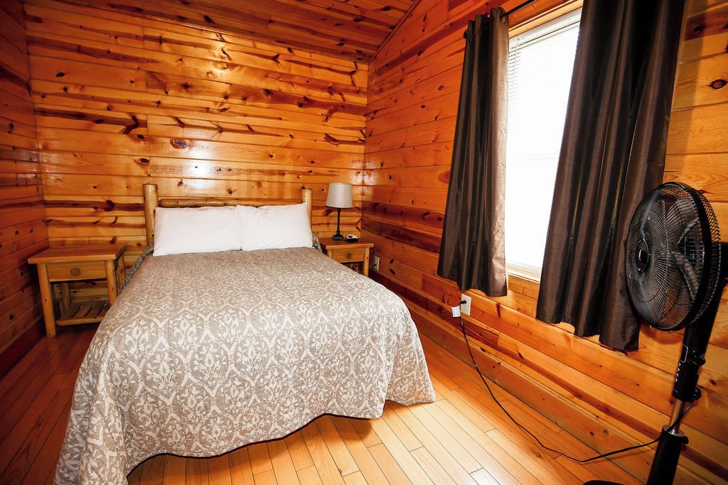 Ein Zimmer in einer Holzhütte mit einem Bett, einem Nachttisch, einer Lampe, einem Fenster mit Vorhängen und einem Standventilator.