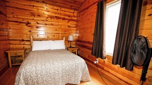 A wooden cabin room with a bed, a nightstand, a lamp, a window with curtains, and a standing fan. - 2 Bedroom 2 Bath Cabin with Hot Tub (Beaver, WV) (Beaver)