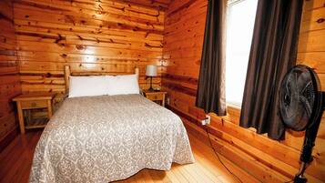Ein Zimmer in einer Holzhütte mit einem Bett, einem Nachttisch, einer Lampe, einem Fenster mit Vorhängen und einem Standventilator.