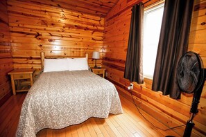 Een houten kamer met een bed, een nachtkastje, een lamp, een raam met gordijnen en een staande ventilator.