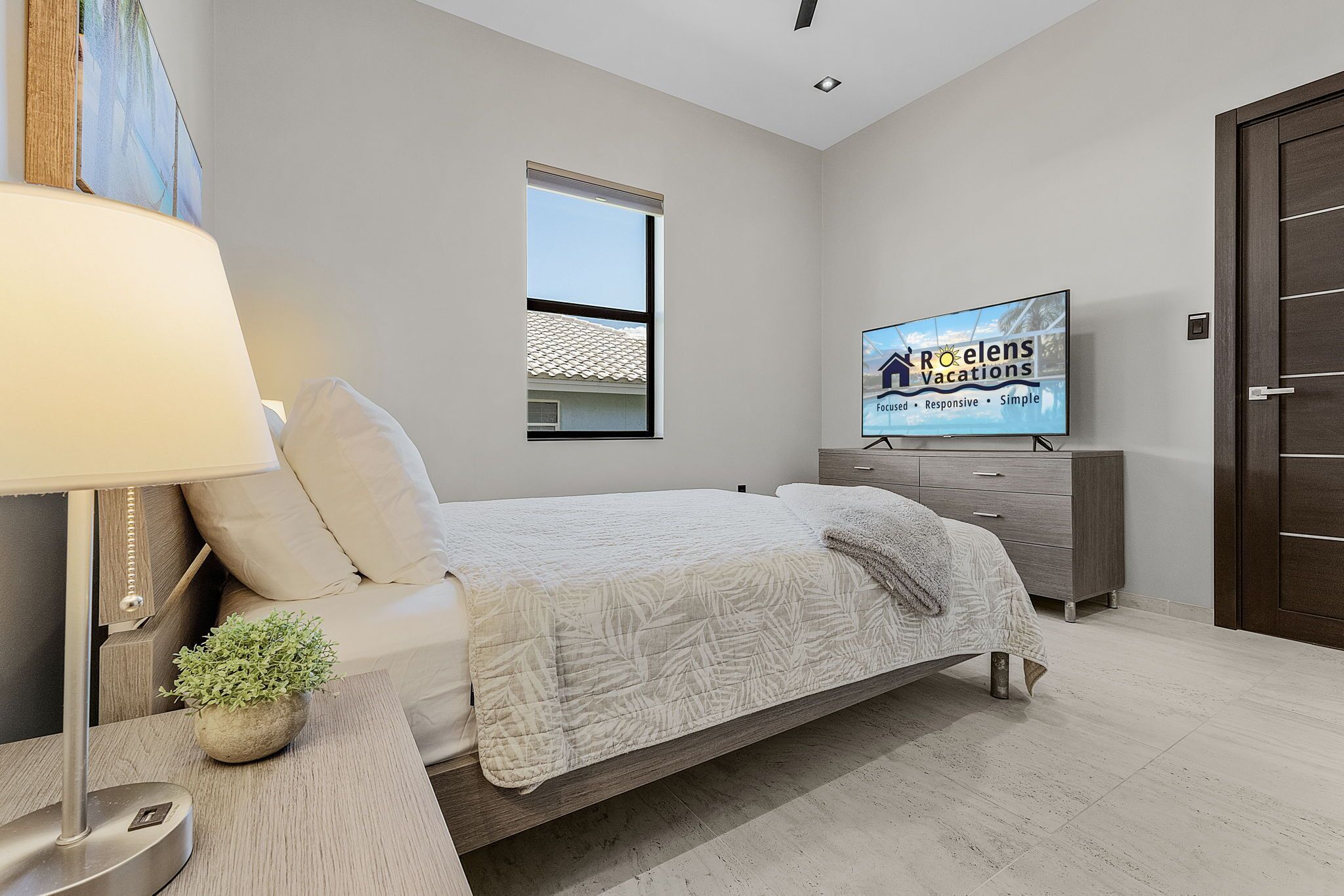 A modern bedroom with a bed, a bedside table with a lamp, a small plant, a TV on the wall, and a window.